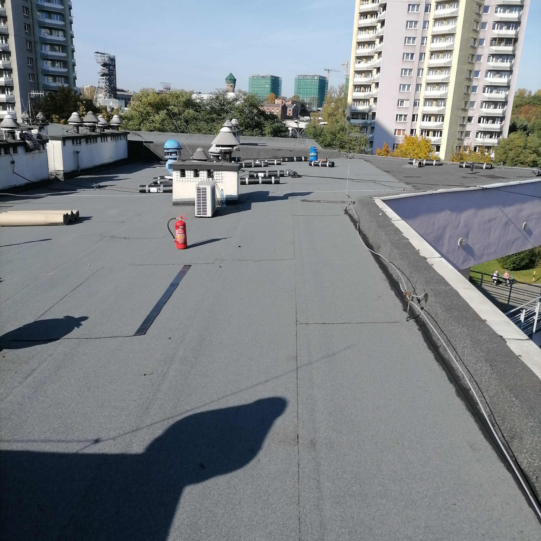 Płaski dach pokryty szarą papą termozgrzewalną z widocznym systemem wentylacji, klimatyzatorem, gaśnicą i zrolowanymi materiałami izolacyjnymi na tle miejskiego krajobrazu