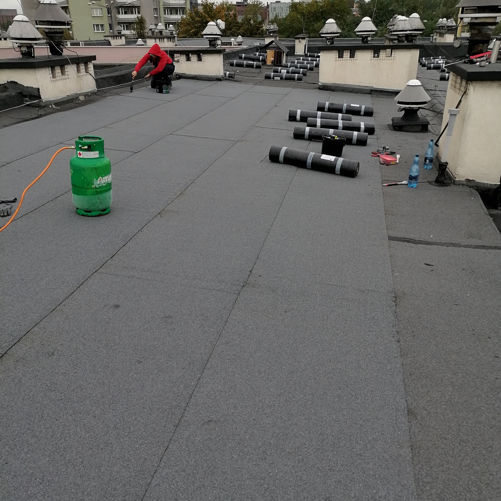 Prace dekarskie w Dąbrowie Górniczej: dekarz w czerwonej bluzie zgrzewa papę na płaskim dachu, obok leżą rolki papy i butla gazowa.