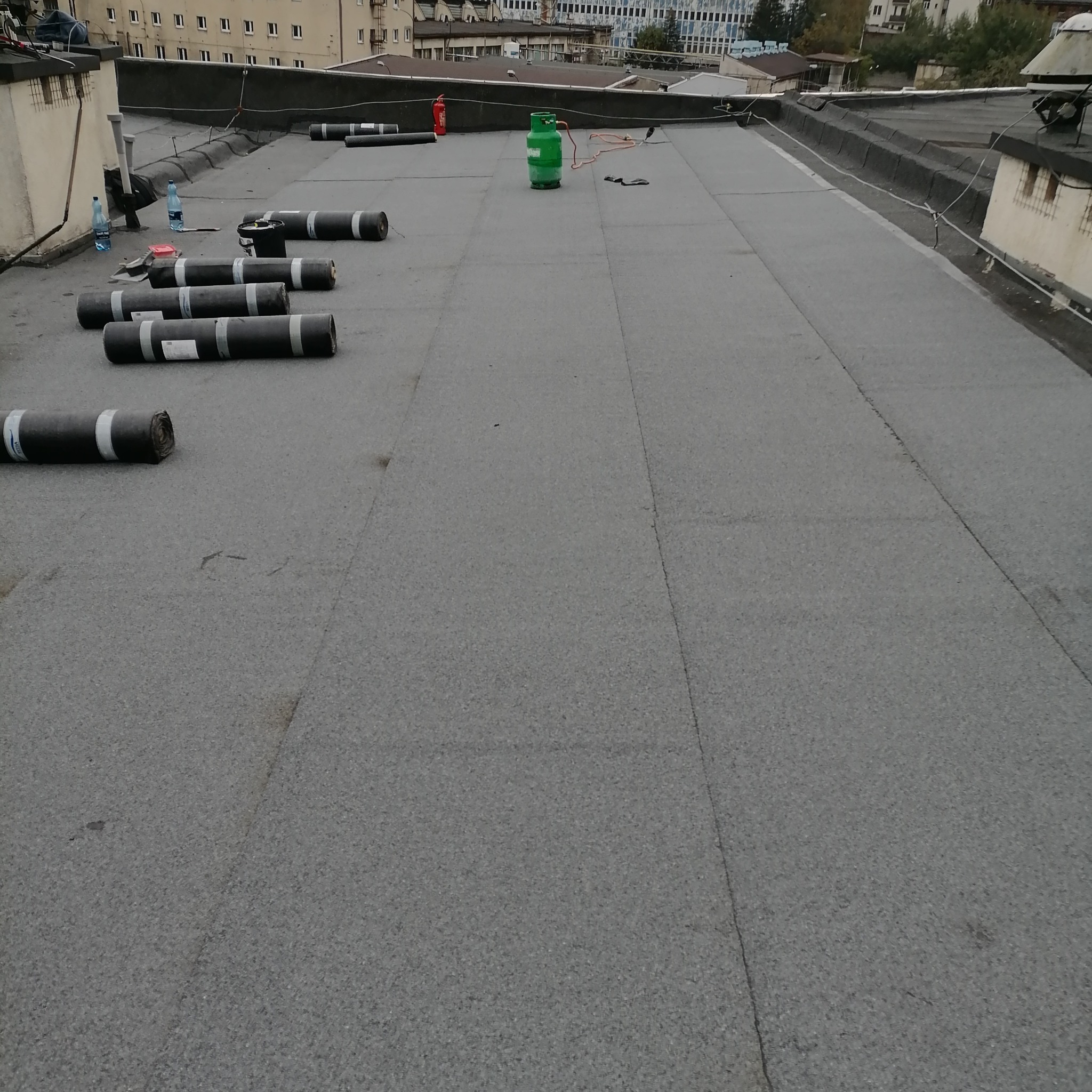 Widok z góry na płaski dach pokryty szarą papą, z rozłożonymi rolkami papy, butlami gazowymi i gaśnicą w tle, sugerujący prace związane z izolacją lub naprawą dachu.