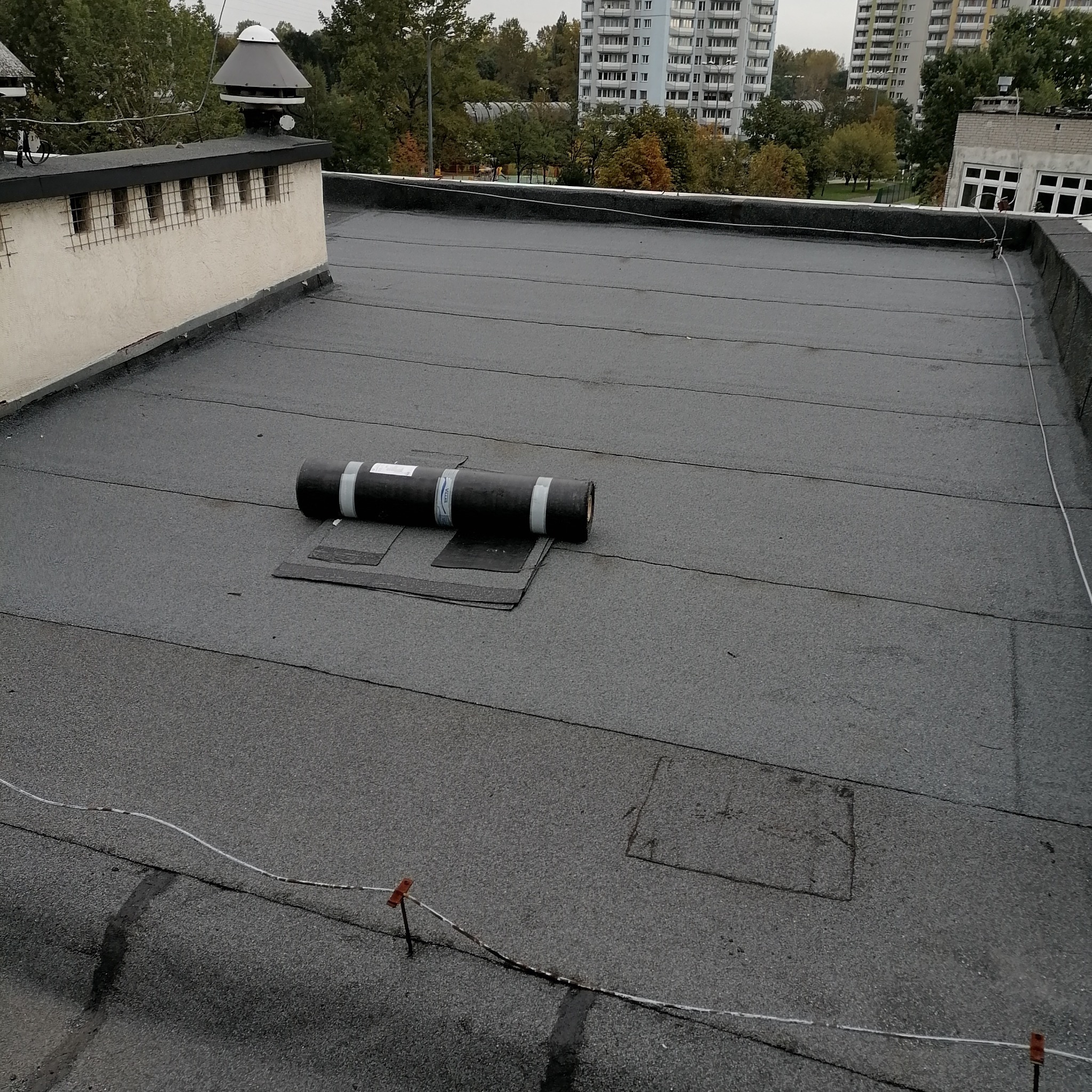 Rolka papy na płaskim dachu pokrytym papą, widoczne zabezpieczenia linką na dachu, w tle budynki mieszkalne i drzewa.