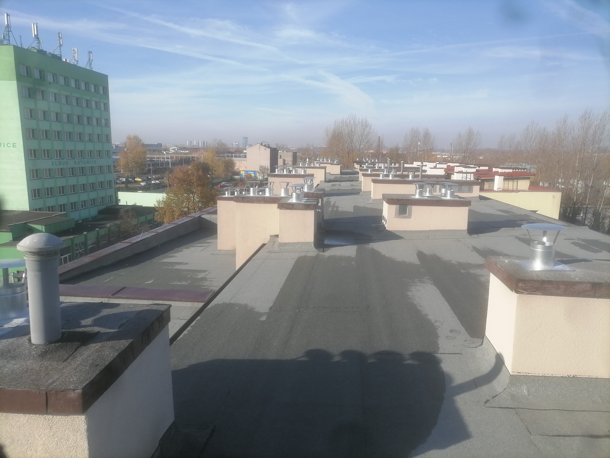 Widok z góry na płaski dach budynku z kominami wentylacyjnymi, w tle budynek biurowy Elbud Katowice i zabudowa miejska w lekkiej mgle.