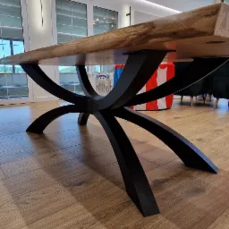 Stół z blatem z litego drewna i czarną, metalową podstawą o oryginalnym, krzyżowym kształcie, widziany z niskiej perspektywy. W tle okna i elementy wystroju wnętrza.