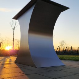 HEFTdesign Paweł Wojewodzic - Metalowa konstrukcja o zakrzywionym kształcie na tle zachodzącego słońca. Element architektoniczny na zewnątrz, widoczny na tle zieleni i ogrodzenia.
