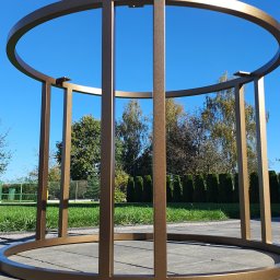 HEFTdesign Paweł Wojewodzic - Metalowa konstrukcja w kolorze złotym na tle niebieskiego nieba i zieleni, prawdopodobnie podstawa stołu lub element dekoracyjny.