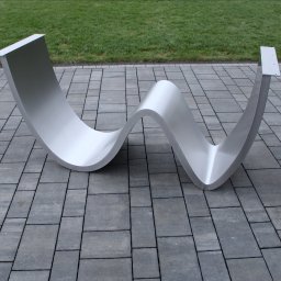 HEFTdesign Paweł Wojewodzic - Falista, nowoczesna konstrukcja ze stali nierdzewnej na szarym bruku, z zielonym trawnikiem w tle. Element architektoniczny o gładkiej powierzchni i zaokrąglonych kształtach.
