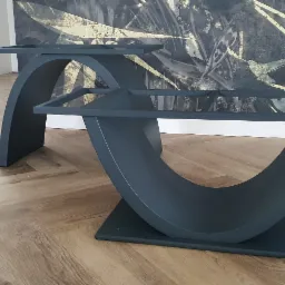 Dwa czarne, metalowe stoliki kawowe o unikalnym kształcie łuku na drewnianej podłodze. W tle ściana z abstrakcyjnym wzorem i białe schody. Nietypowy design.