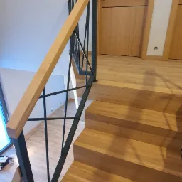 Drewniane schody z czarną, geometryczną balustradą i drewnianą poręczą, widok z góry, drzwi z forniru w tle.