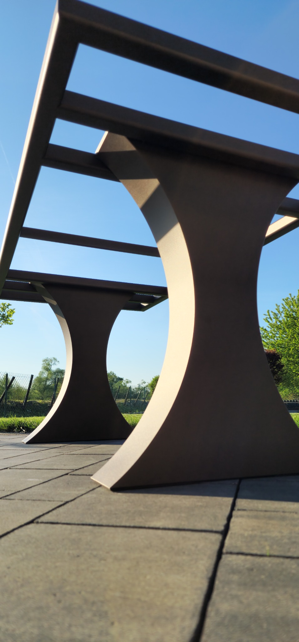 Metalowa konstrukcja stołu ogrodowego z charakterystycznymi, półokrągłymi nogami, widziana z niskiej perspektywy na tle błękitnego nieba i zieleni.