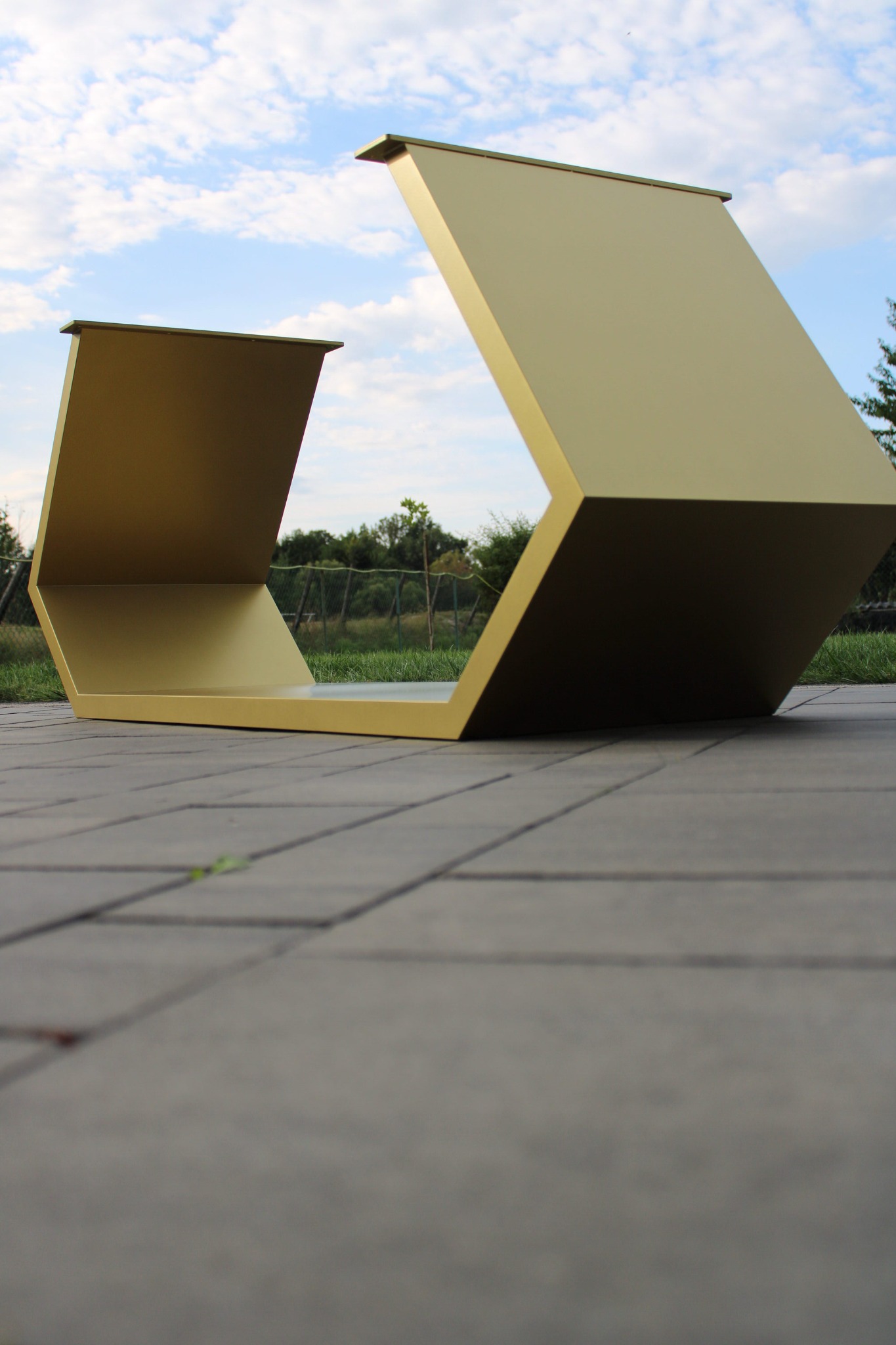 Nowoczesna, złota konstrukcja metalowa w kształcie litery U, ustawiona na szarym bruku. Tło stanowią zieleń i błękitne niebo z chmurami. Niska perspektywa.