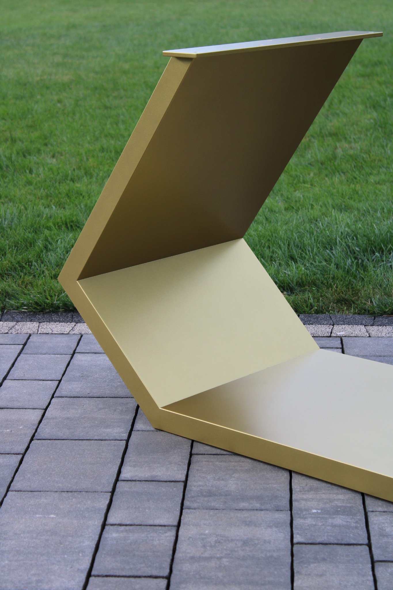 Złota, geometryczna konstrukcja metalowa na szarym bruku i tle zielonego trawnika. Awangardowa forma, precyzyjne wykonanie, minimalistyczny design.