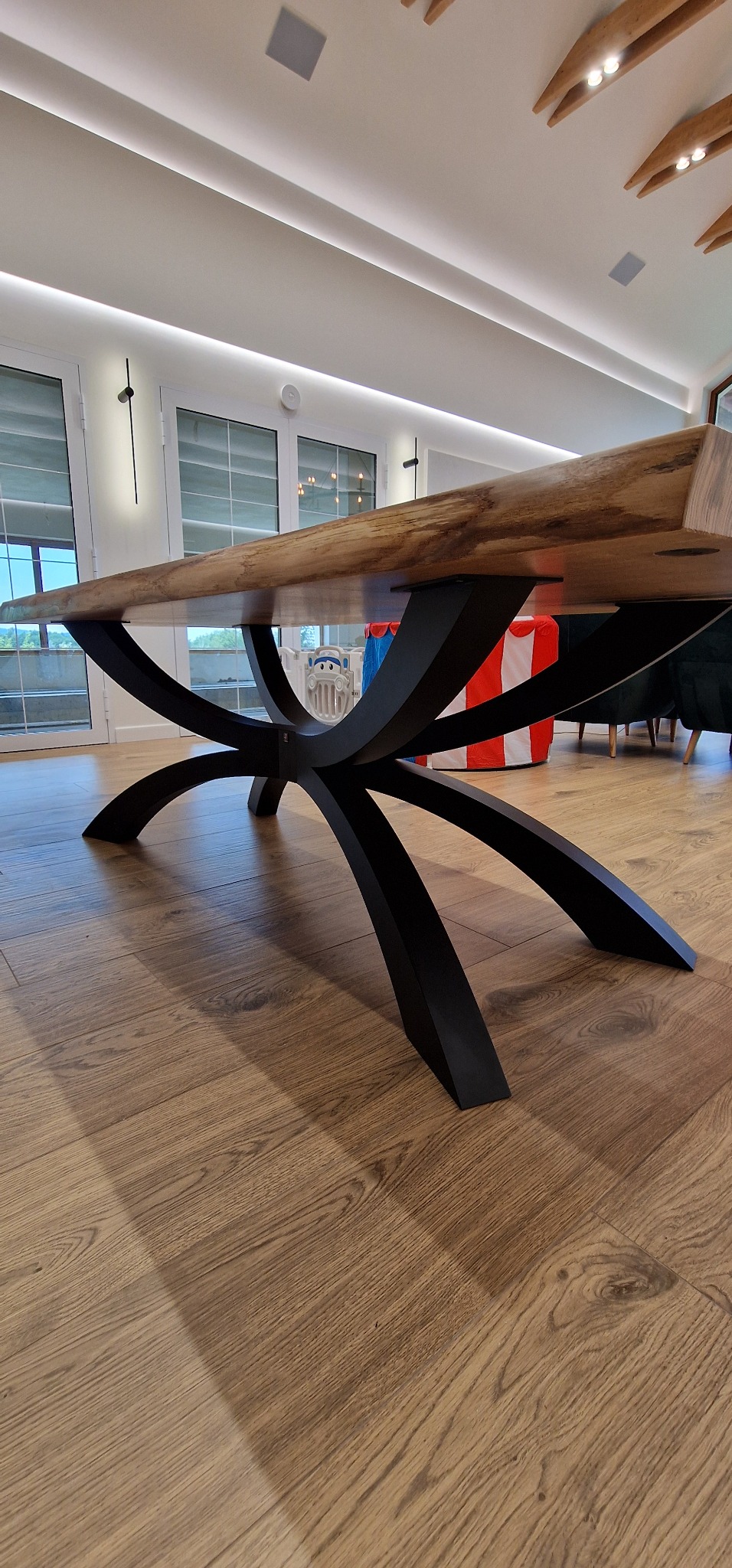 Stół z blatem z litego drewna i czarną, metalową podstawą o oryginalnym, krzyżowym kształcie, widziany z niskiej perspektywy. W tle okna i elementy wystroju wnętrza.