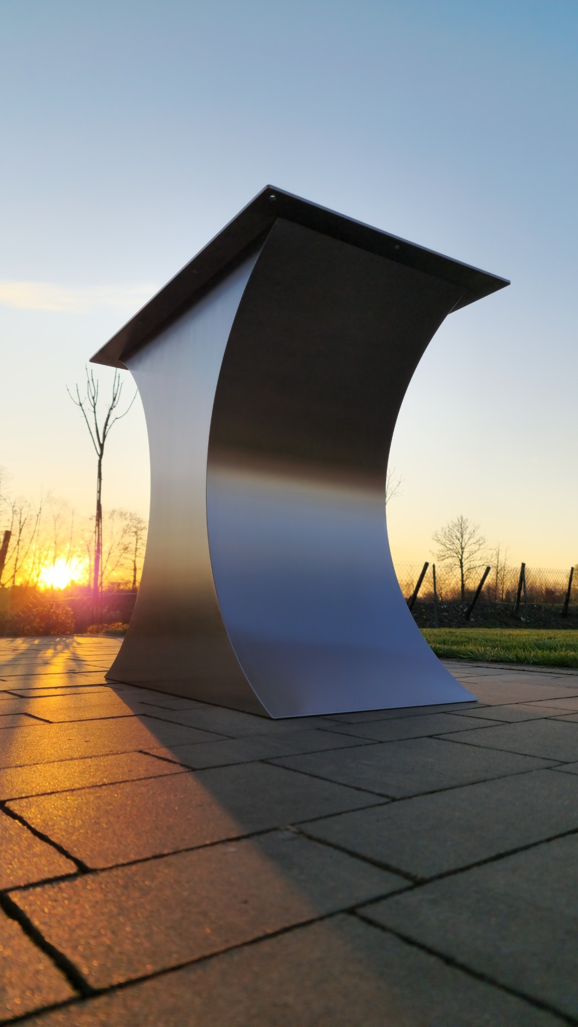 Metalowa konstrukcja o zakrzywionym kształcie na tle zachodzącego słońca. Element architektoniczny na zewnątrz, widoczny na tle zieleni i ogrodzenia.