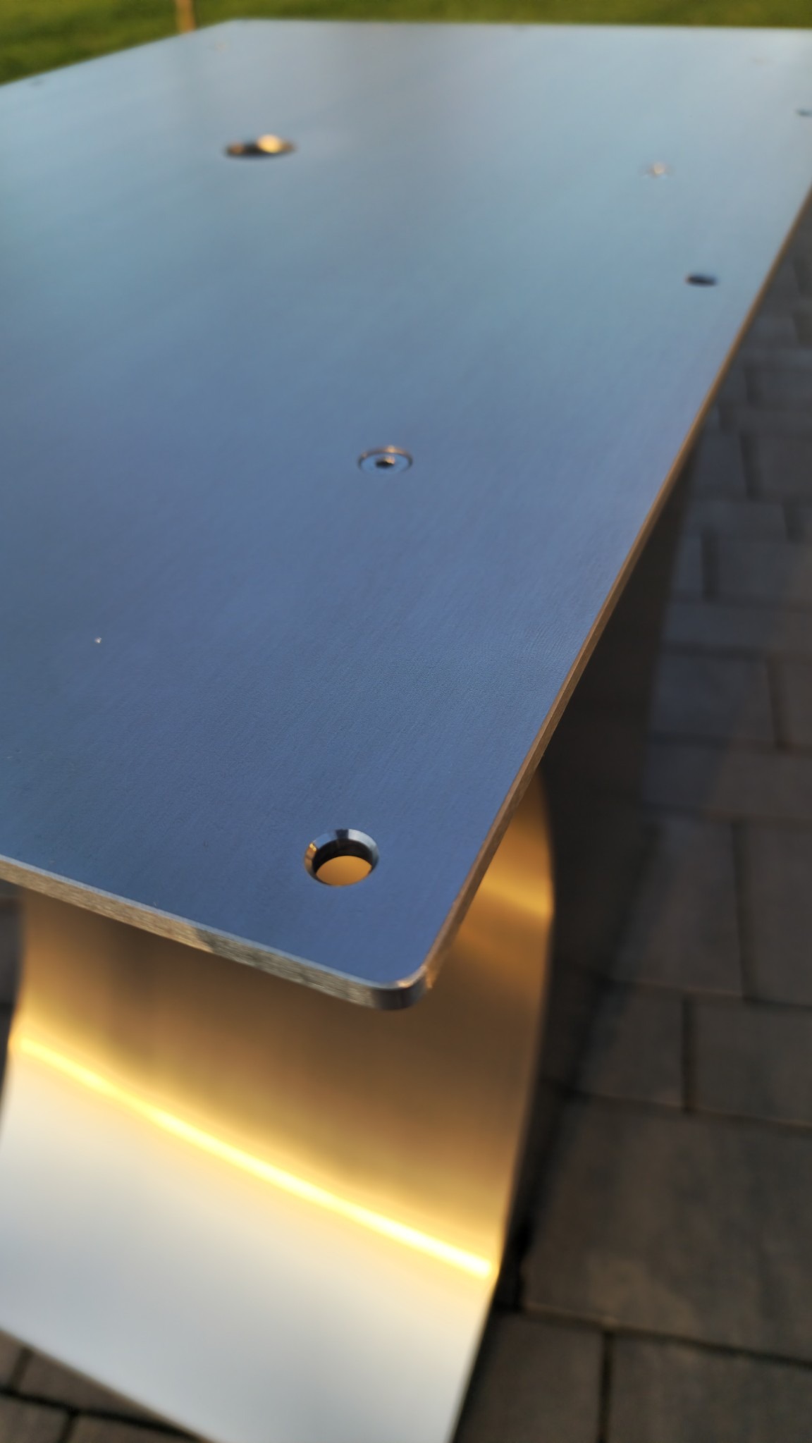 Detal metalowego stolika z blatem z otworami, na tle kostki brukowej. Oświetlenie podkreśla gładką powierzchnię stali.