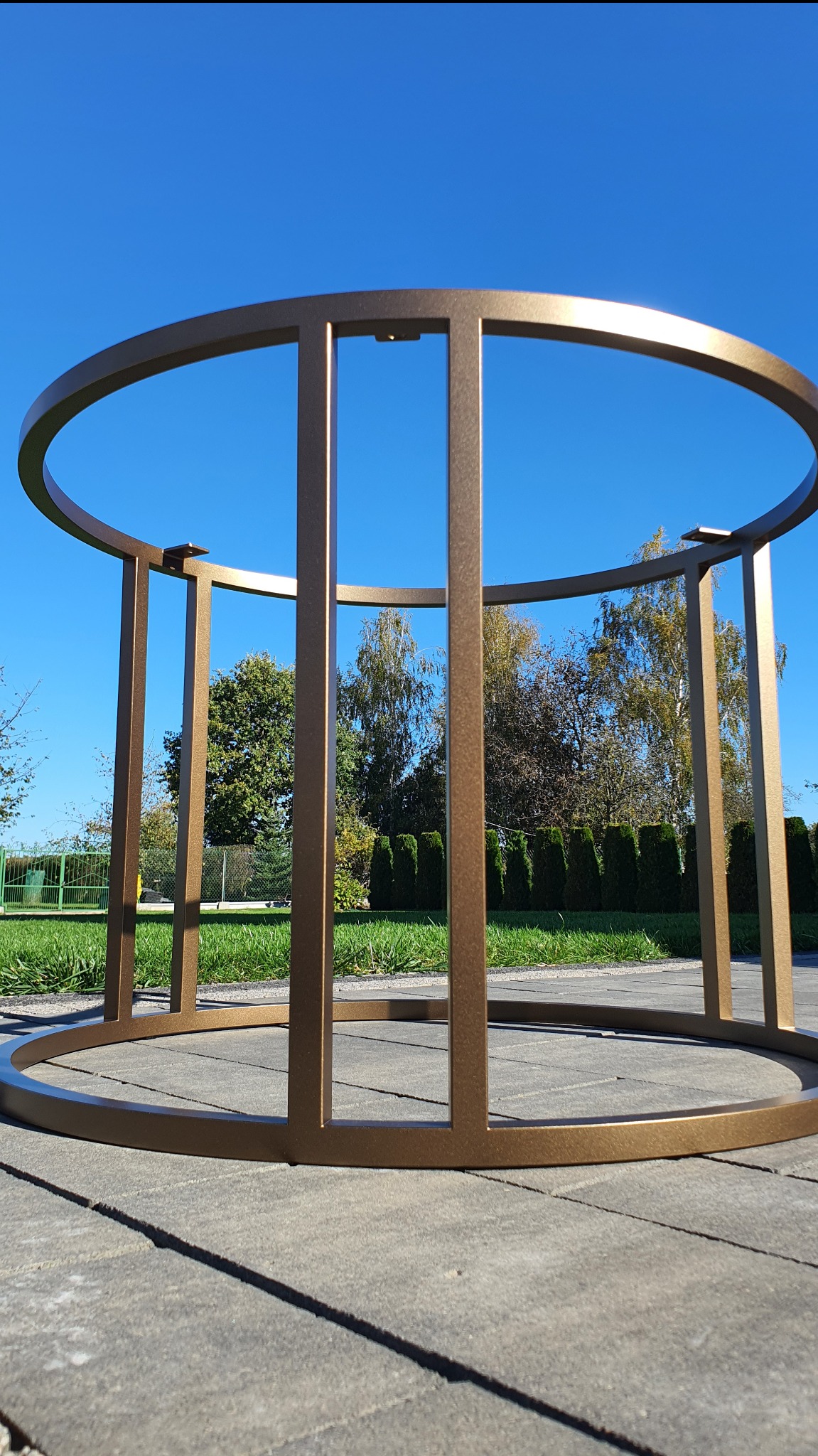 Metalowa konstrukcja w kolorze złotym na tle niebieskiego nieba i zieleni, prawdopodobnie podstawa stołu lub element dekoracyjny.