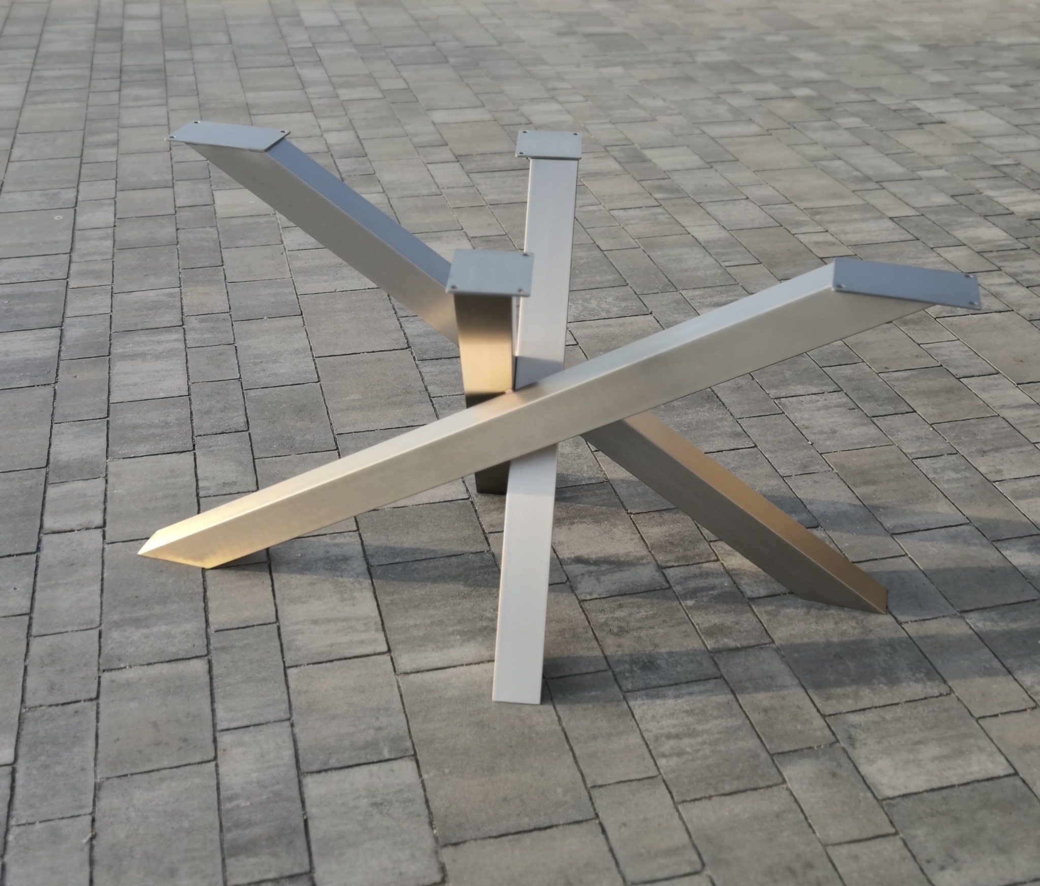 Nowoczesna, geometryczna podstawa stołu z metalu, z trzema ramionami krzyżującymi się pod kątem. Wykończenie w odcieniach szarości i srebra, na tle szarej kostki brukowej.