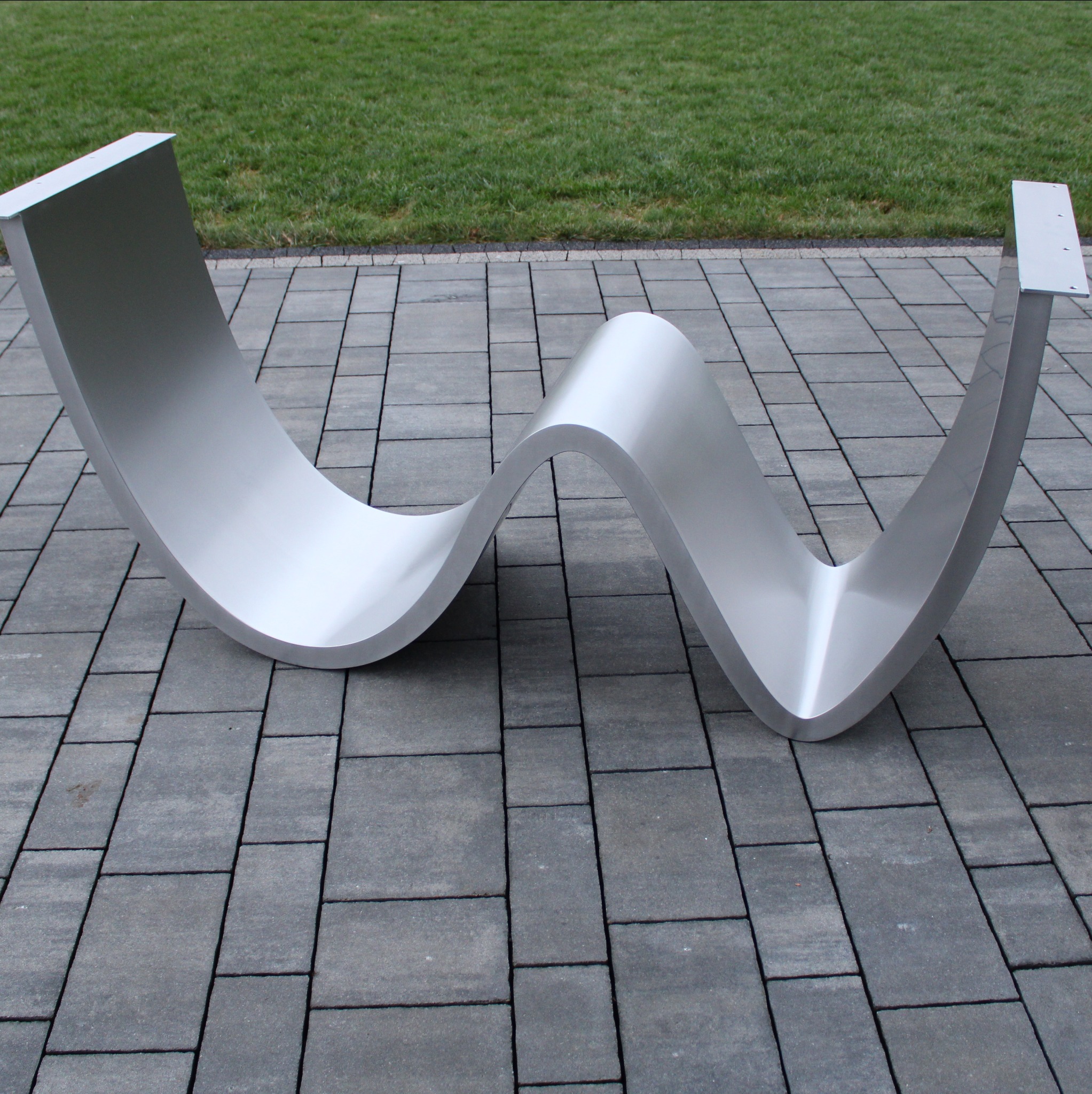 Falista, nowoczesna konstrukcja ze stali nierdzewnej na szarym bruku, z zielonym trawnikiem w tle. Element architektoniczny o gładkiej powierzchni i zaokrąglonych kształtach.