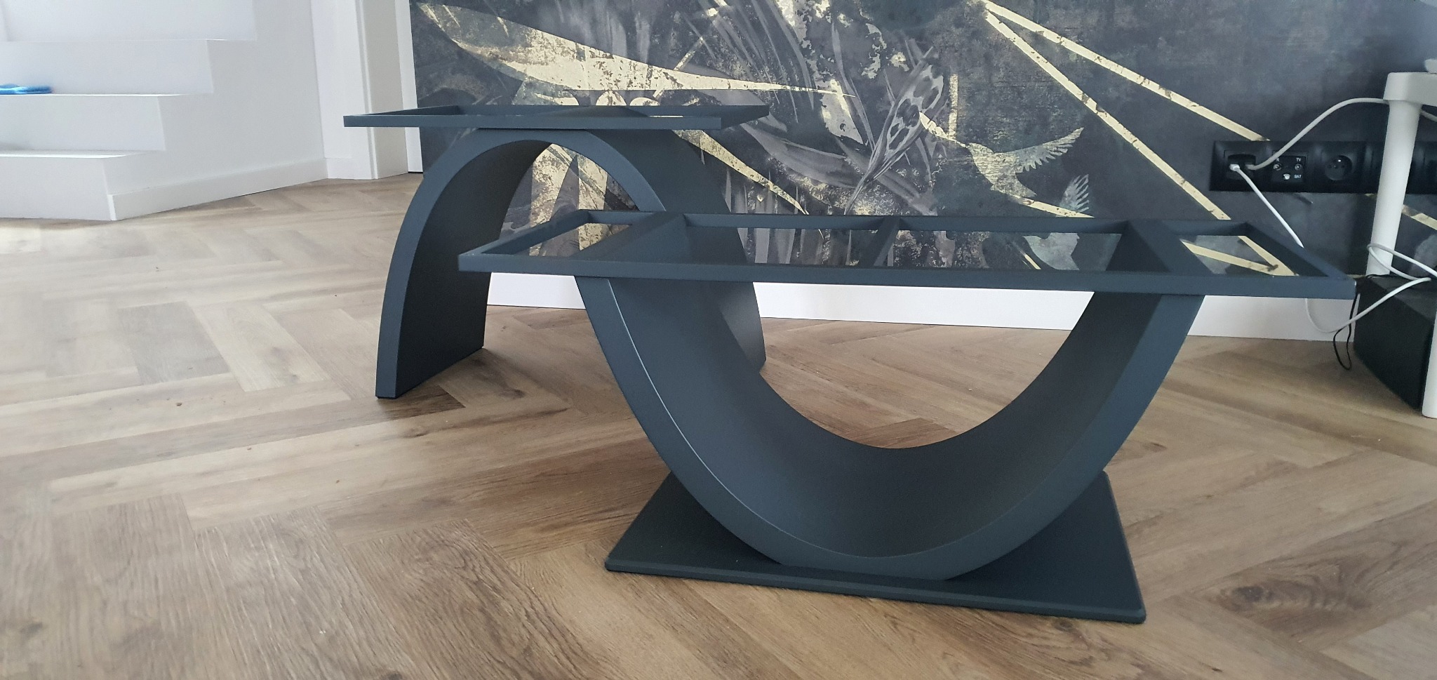 Dwa czarne, metalowe stoliki kawowe o unikalnym kształcie łuku na drewnianej podłodze. W tle ściana z abstrakcyjnym wzorem i białe schody. Nietypowy design.