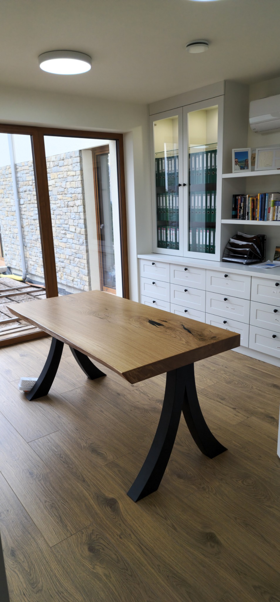 Stół dębowy z czarnymi, metalowymi nogami w kształcie litery X, na tle jasnego wnętrza z dużym oknem i zabudową meblową.