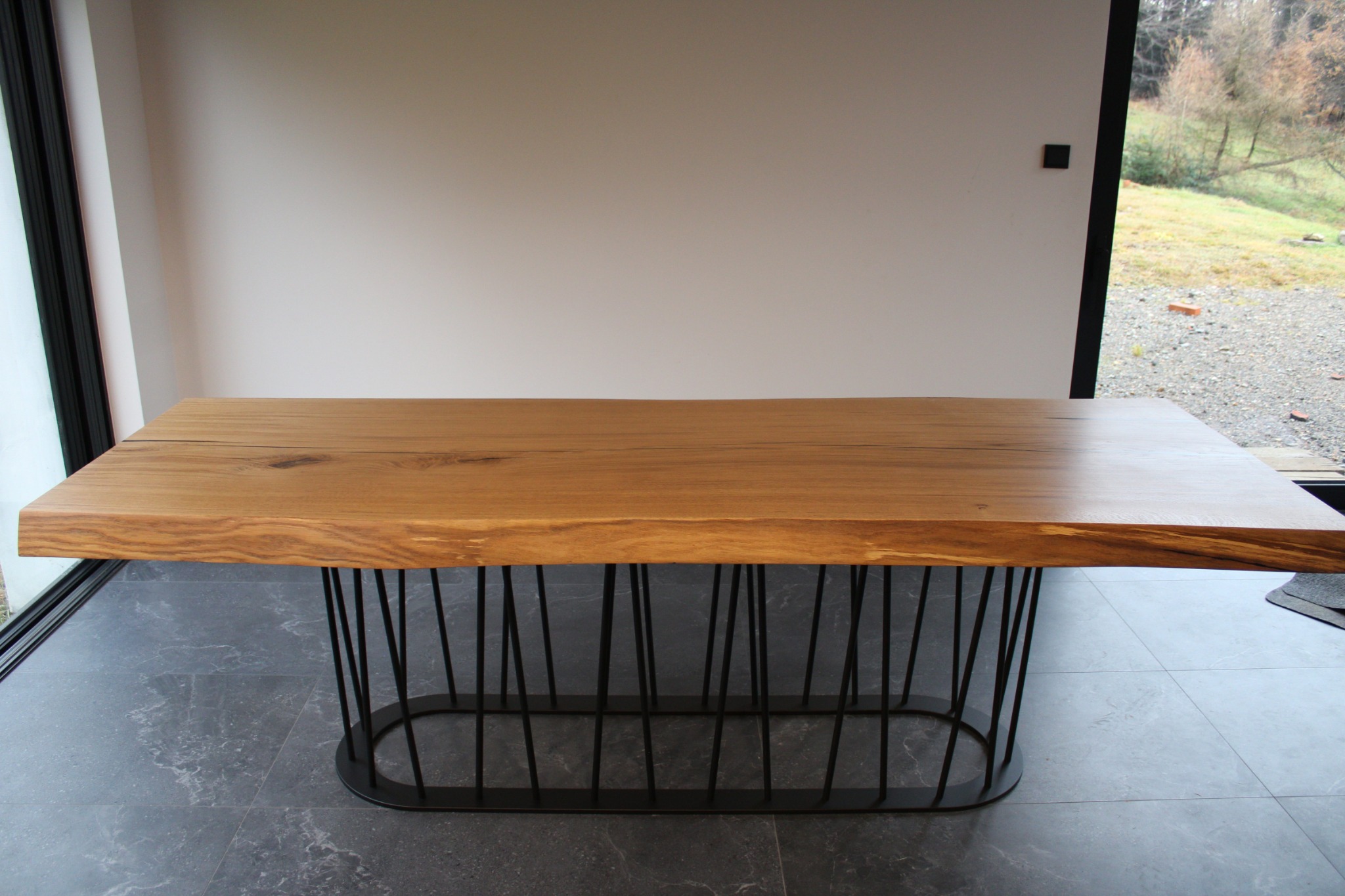Stół z blatem z litego drewna o nieregularnej krawędzi, wsparty na metalowej podstawie z prętów. Nowoczesny design, jasne wnętrze, widok na zewnątrz.