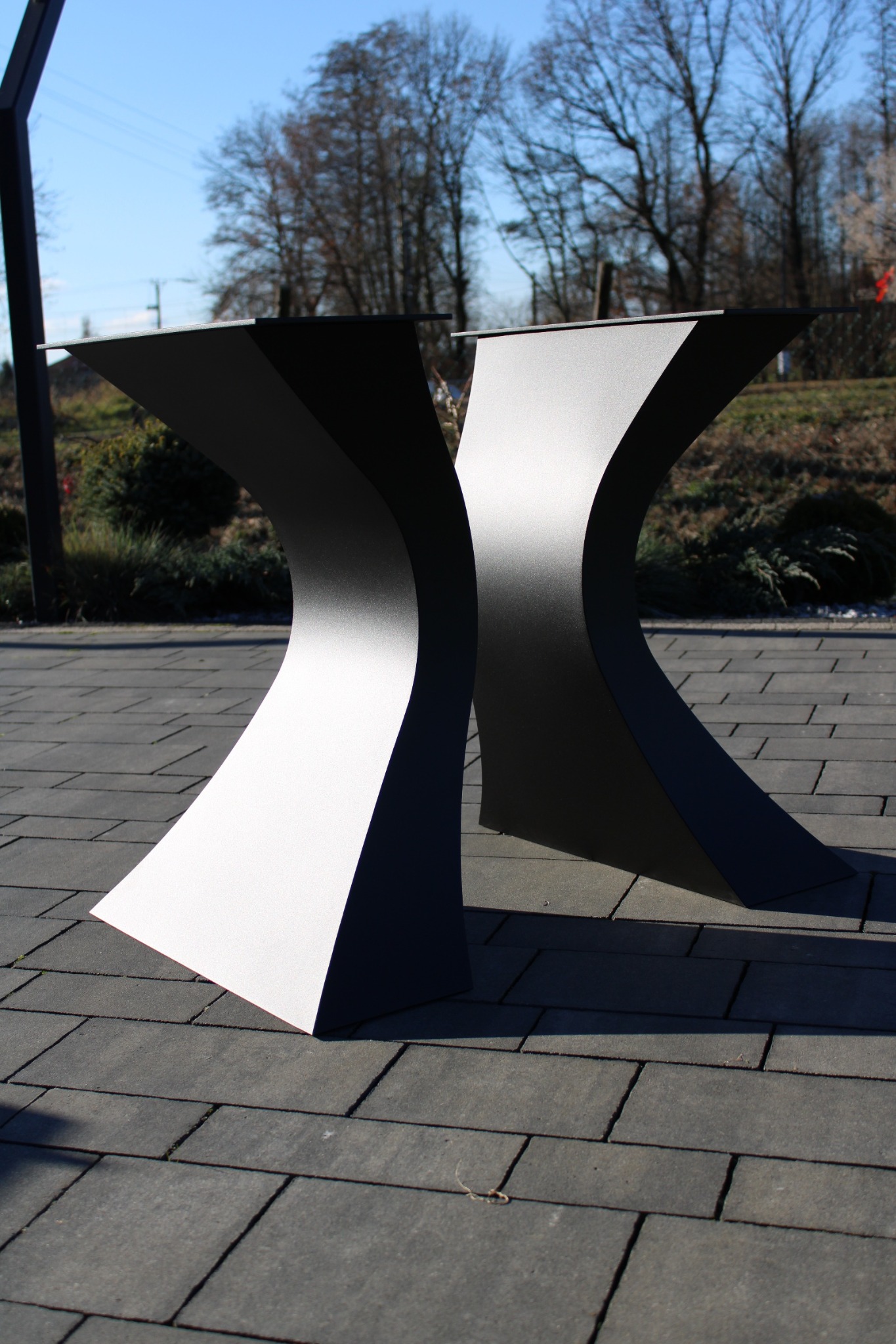 Nowoczesny, metalowy stół ogrodowy z oryginalną podstawą w kształcie klepsydry, ustawiony na szarej kostce brukowej. Minimalistyczny design, błękitne niebo w tle.