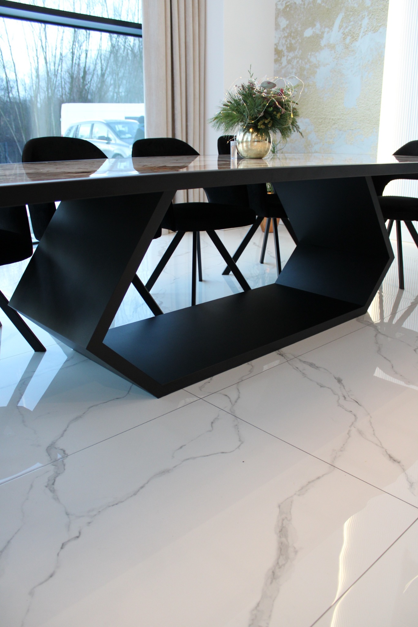 Stół z blatem imitującym drewno i geometryczną, czarną podstawą. Na stole dekoracja roślinna w złotej kuli. Białe, marmurowe płytki podłogowe.