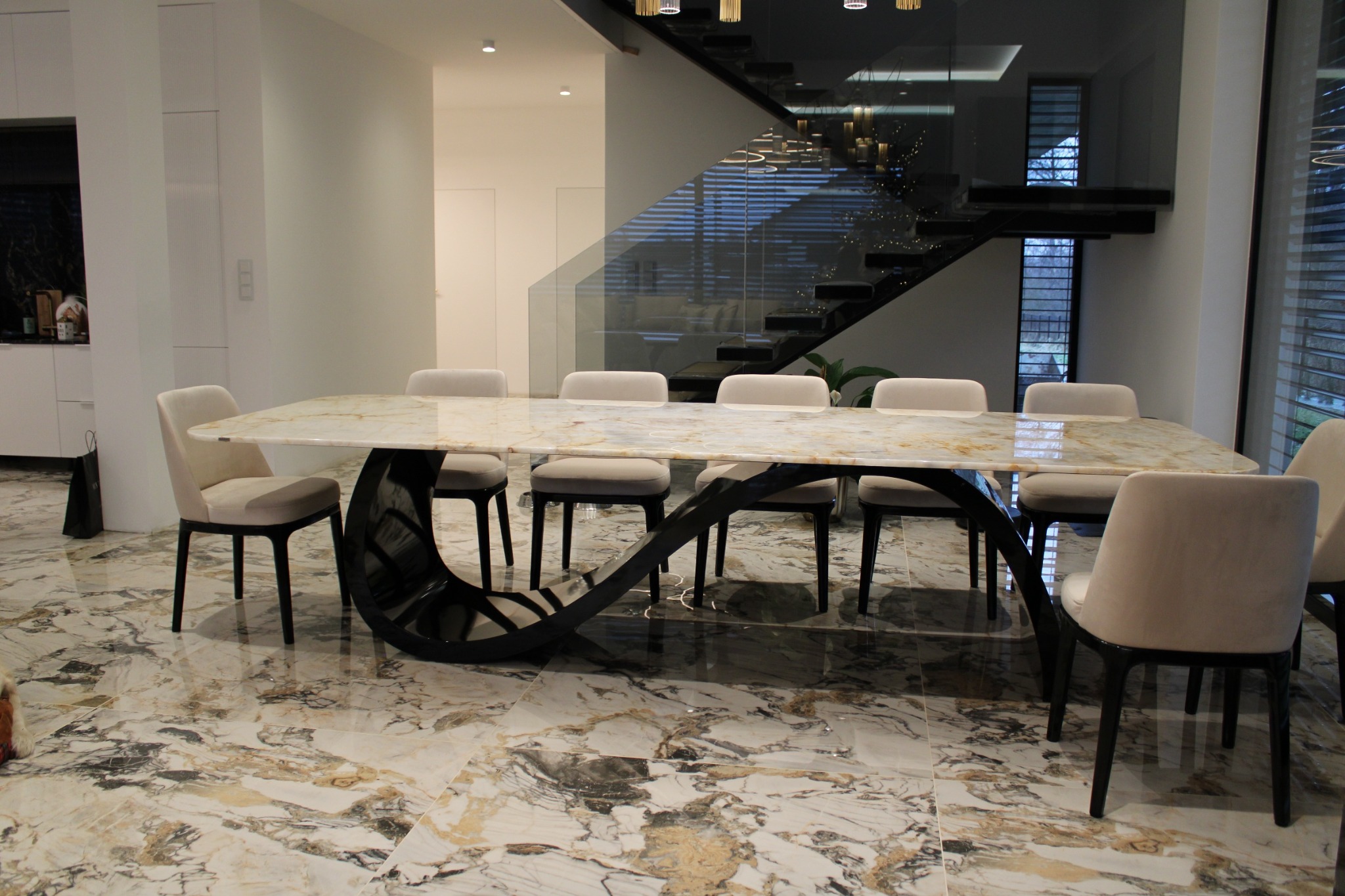 Stylowa jadalnia z marmurowym stołem na czarnej, designerskiej podstawie. W tle schody z szklaną balustradą i nowoczesne oświetlenie.