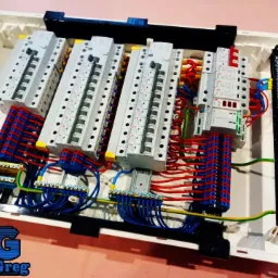 Rozdzielnica elektryczna z czterema rzędami zabezpieczeń, okablowana przewodami w kolorach niebieskim, czerwonym i żółto-zielonym, widoczny logo firmy VoltGreg w lewym dolnym rogu.