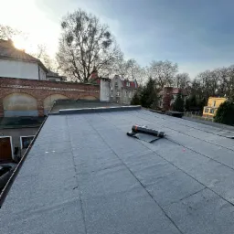 Widok z góry na płaski dach pokryty papą, z rolką papy i narzędziem do zgrzewania leżącymi na powierzchni, w tle budynki mieszkalne i drzewa bez liści pod błękitnym niebem.