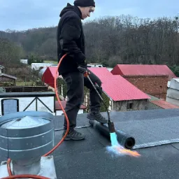 Pracownik w czarnym ubraniu z palnikiem na dachu, termozgrzewalna papa w trakcie montażu, widoczny płomień palnika i struktura papy, w tle zabudowania i drzewa.