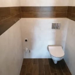 Nowoczesna toaleta podwieszana w wykończonej łazience z brązowymi płytkami imitującymi drewno na podłodze i pasem na ścianie, oraz jasnymi płytkami imitującymi kamień na pozostałej powierzchni ścian.