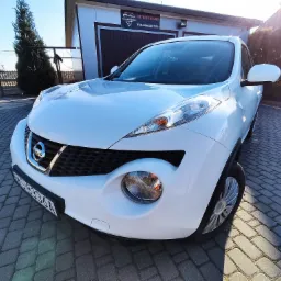 Biały Nissan Juke zaparkowany na kostce brukowej przed budynkiem z szyldem 'Autodetailing'.