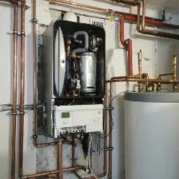 Nowoczesny piec centralnego ogrzewania z otwartą obudową, widoczne wewnętrzne komponenty, podłączony do miedzianych rur w piwnicy z surowymi ścianami.