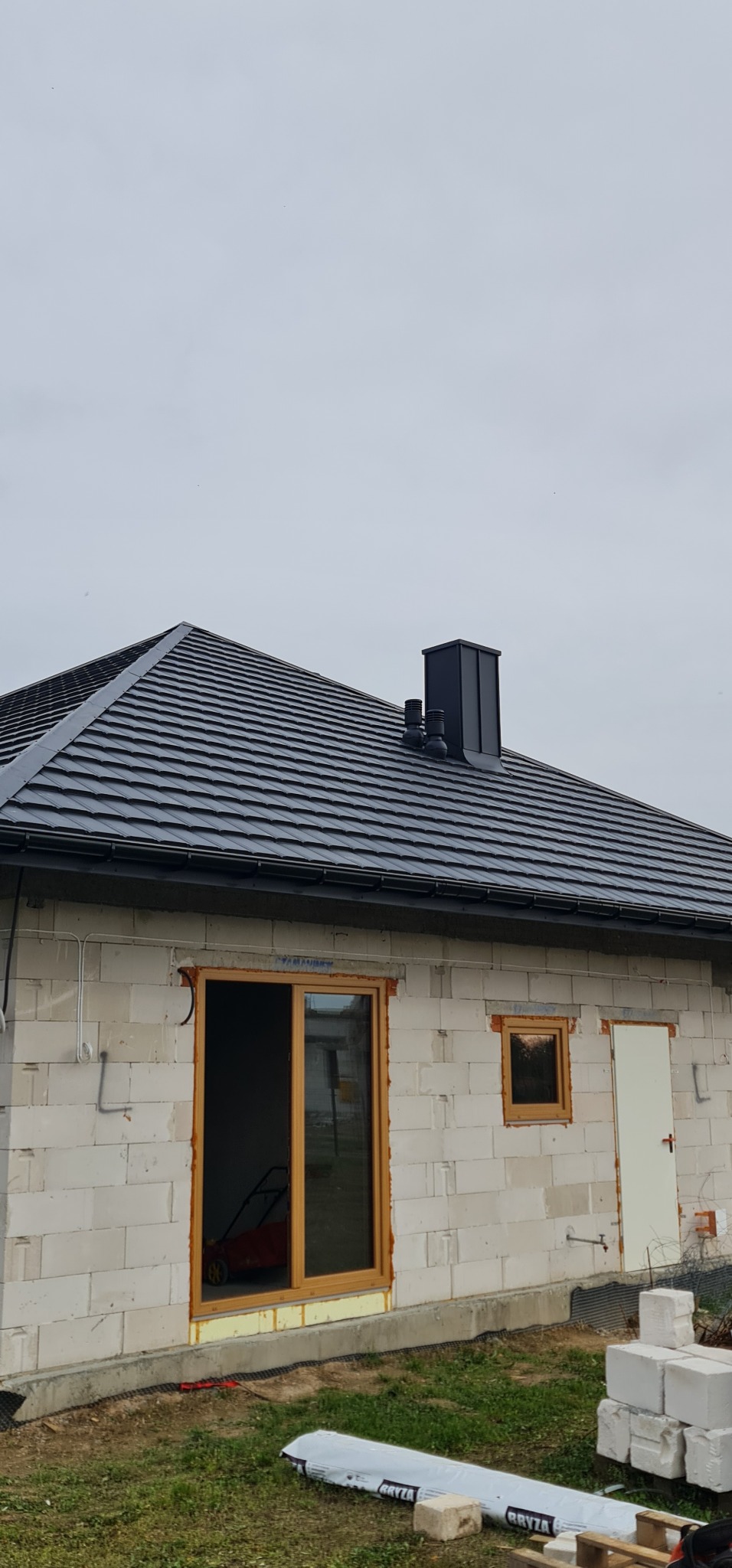 Dom w Sompolnie w trakcie budowy, widoczny nowy dach z czarnej dachówki, komin i ściany z białego bloczka, okna z drewnianą ramą oraz materiały budowlane na trawniku.