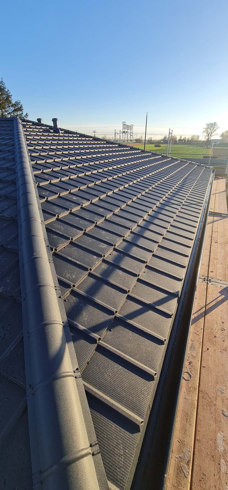 Nowy dach z szarej blachodachówki modułowej, widok z góry na całą połać z zamontowanym orynnowaniem, w tle błękitne niebo i słupy wysokiego napięcia.