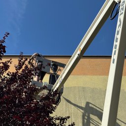 AML Company - Pracownik na podnośniku koszowym czyści elewację budynku o pastelowych pasach, widok z dołu, z krzewem o bordowych liściach w kadrze.