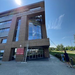 AML Company - Mycie okien Starostwa Powiatowego w Kielcach. Pracownik używa teleskopowej lancy. Obok budynku stoi drabina. Błękitne niebo z cirrus fibratus w tle.