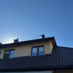Pracownik w kasku czyści ciemny dach domu jednorodzinnego za pomocą lancy teleskopowej, na tle błękitnego nieba.