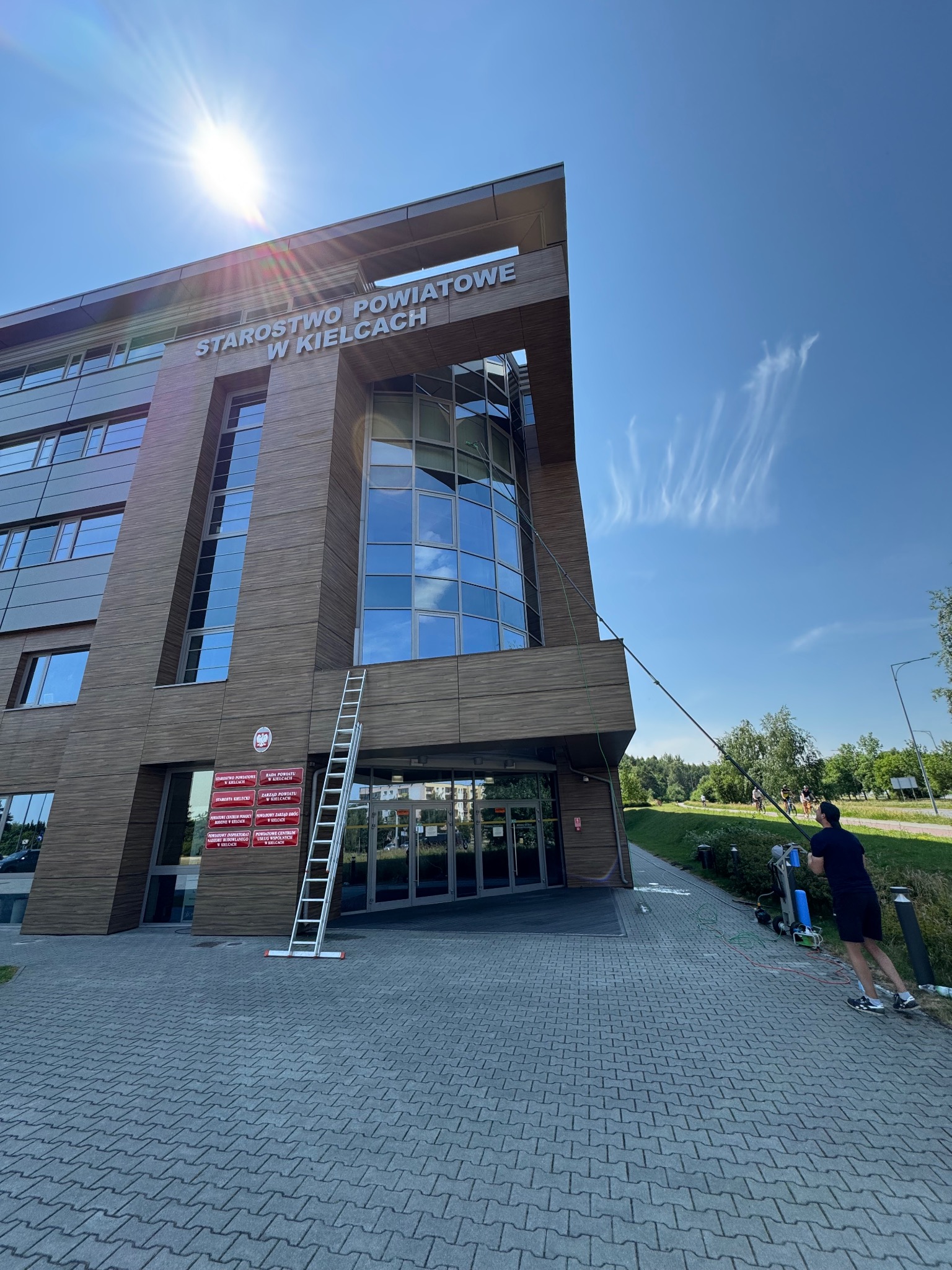 Mycie okien Starostwa Powiatowego w Kielcach. Pracownik używa teleskopowej lancy. Obok budynku stoi drabina. Błękitne niebo z cirrus fibratus w tle.