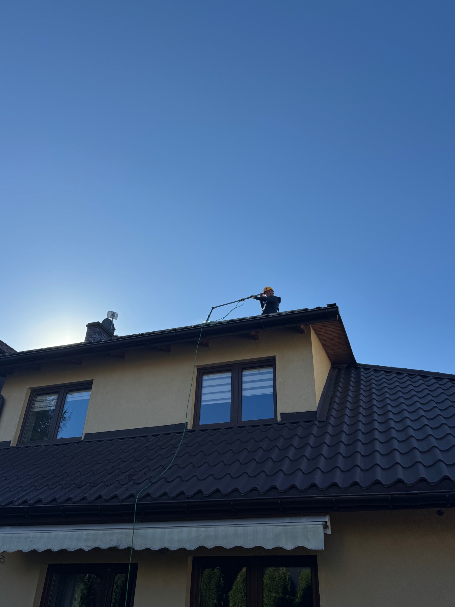 Pracownik w kasku czyści ciemny dach domu jednorodzinnego za pomocą lancy teleskopowej, na tle błękitnego nieba.