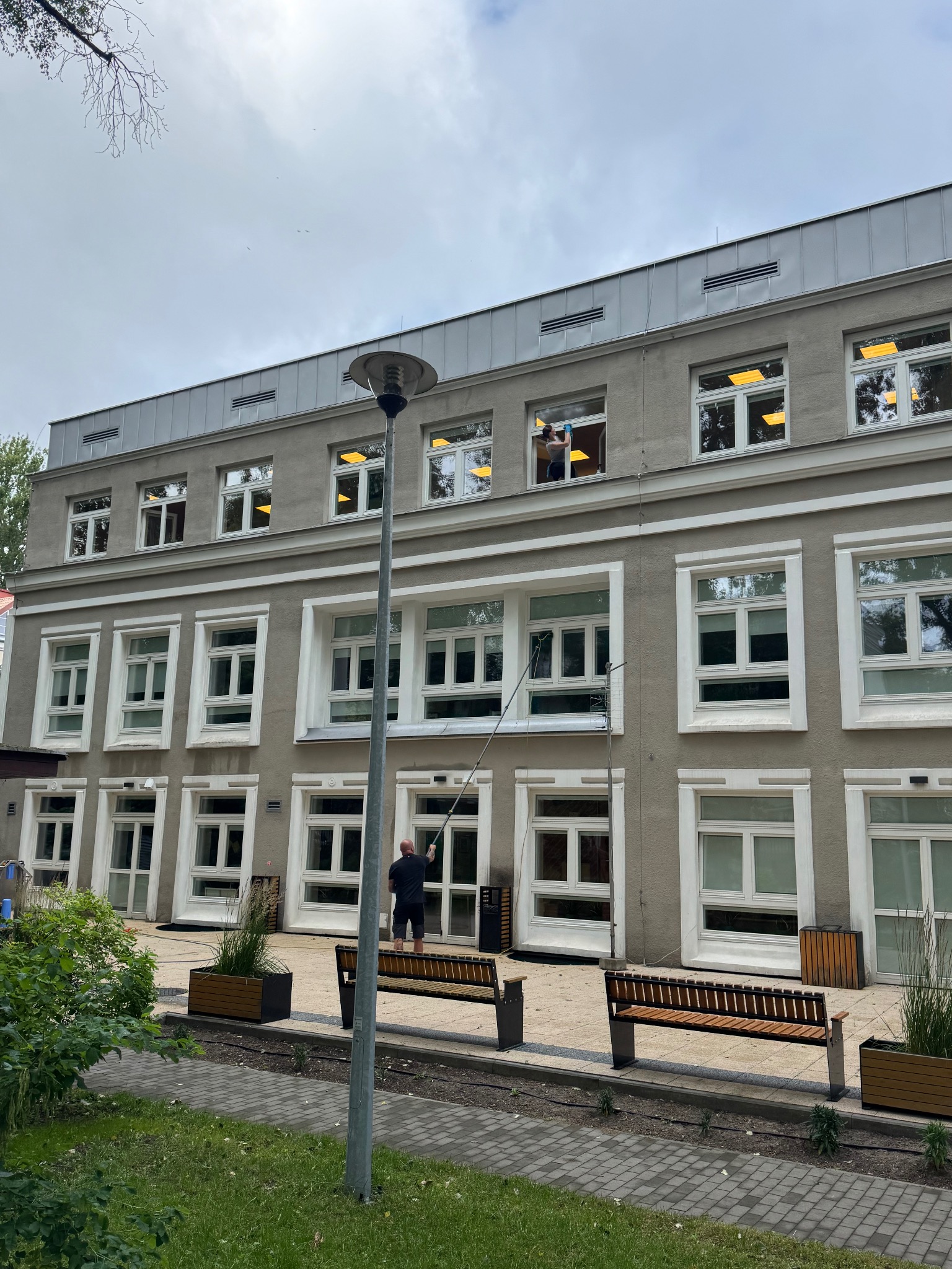 Pracownik myje okna budynku z zewnątrz za pomocą długiej lancy. Widoczne ławki i kwietniki przed budynkiem, pochmurne niebo w tle. Dzień pracy w toku.