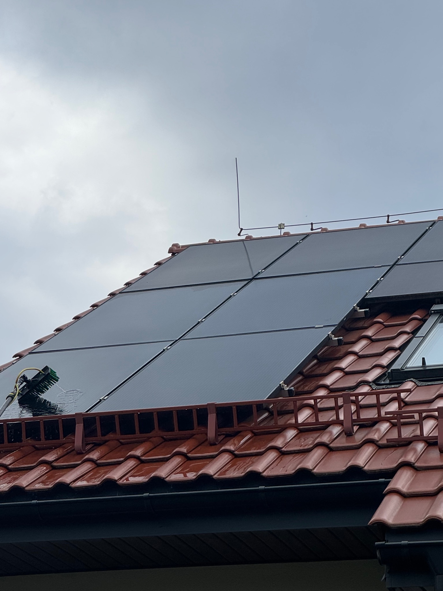 Panel fotowoltaiczny na dachu z czerwoną dachówką, czyszczony szczotką z wodą. Widoczny fragment okna dachowego i pochmurne niebo.