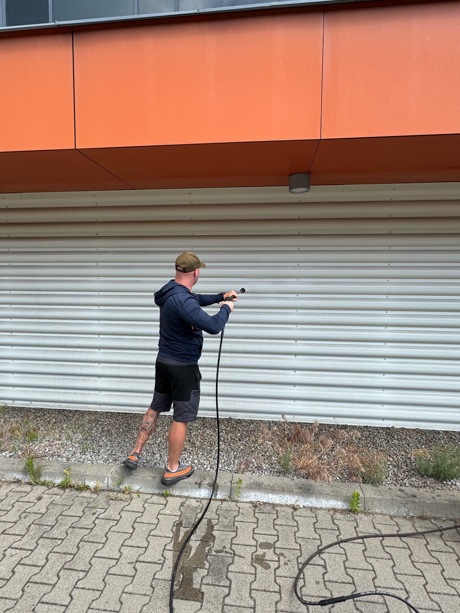 Pracownik myje ciśnieniowo metalową elewację budynku z pomarańczowymi panelami. Widoczny wąż i dysza spryskująca ścianę. Czyszczenie elewacji w toku.