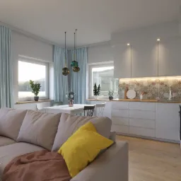 Jasne wnętrze salonu z aneksem kuchennym; sofa z ż&oacute;łtą poduszką na pierwszym planie, st&oacute;ł jadalny i kuchnia z mozaiką na ścianie w tle.