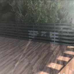 Szare, metalowe balustrady balkonowe z poziomymi szczeblami, na drewnopodobnej podłodze tarasu, z zielenią w tle.