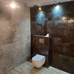 Nowoczesna łazienka z płytkami imitującymi rdzę na ścianie, podwieszana toaleta z chromowanym przyciskiem spłukującym, oraz szare płytki na ścianach i podłodze, oświetlona punktowymi lampami.