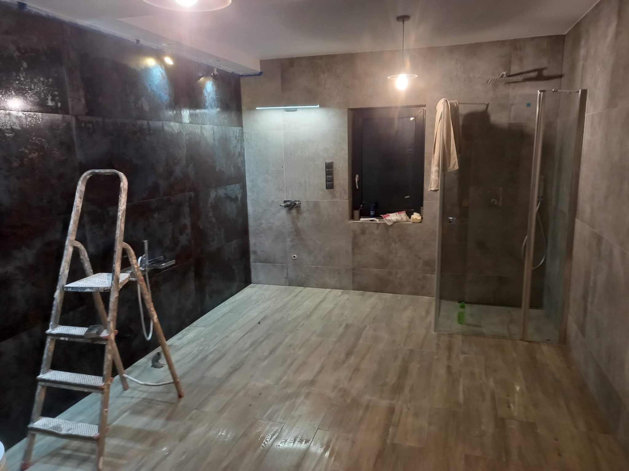 Łazienka w trakcie remontu: ciemne płytki na ścianach, jasne na podłodze, drabina malarska obok ściany, kabina prysznicowa w rogu.