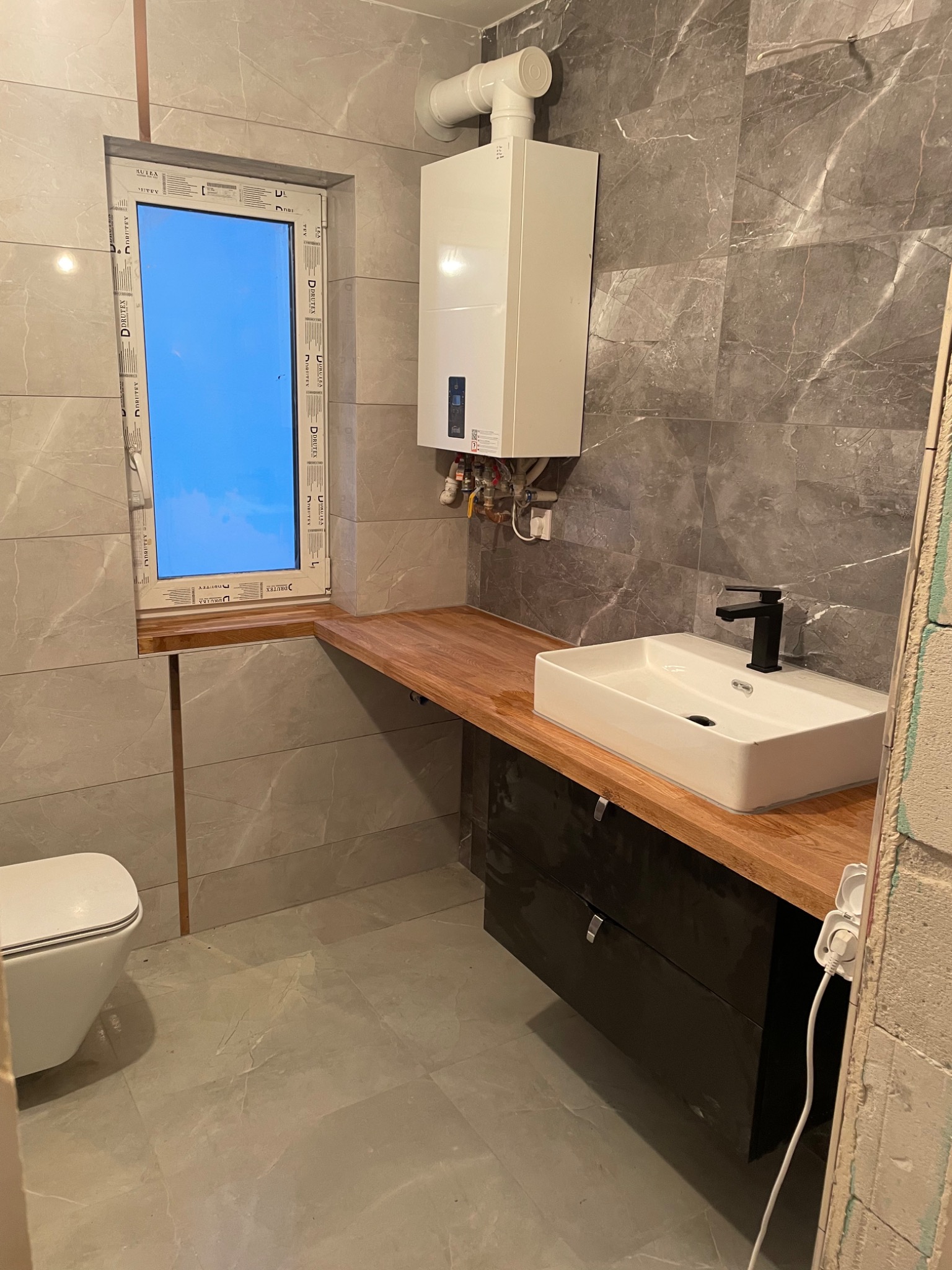 Nowoczesna łazienka w trakcie wykańczania: szare płytki imitujące marmur, biała umywalka nablatowa z czarną armaturą na drewnianym blacie z czarną szafką, wiszący piec gazowy, okno z widokiem...