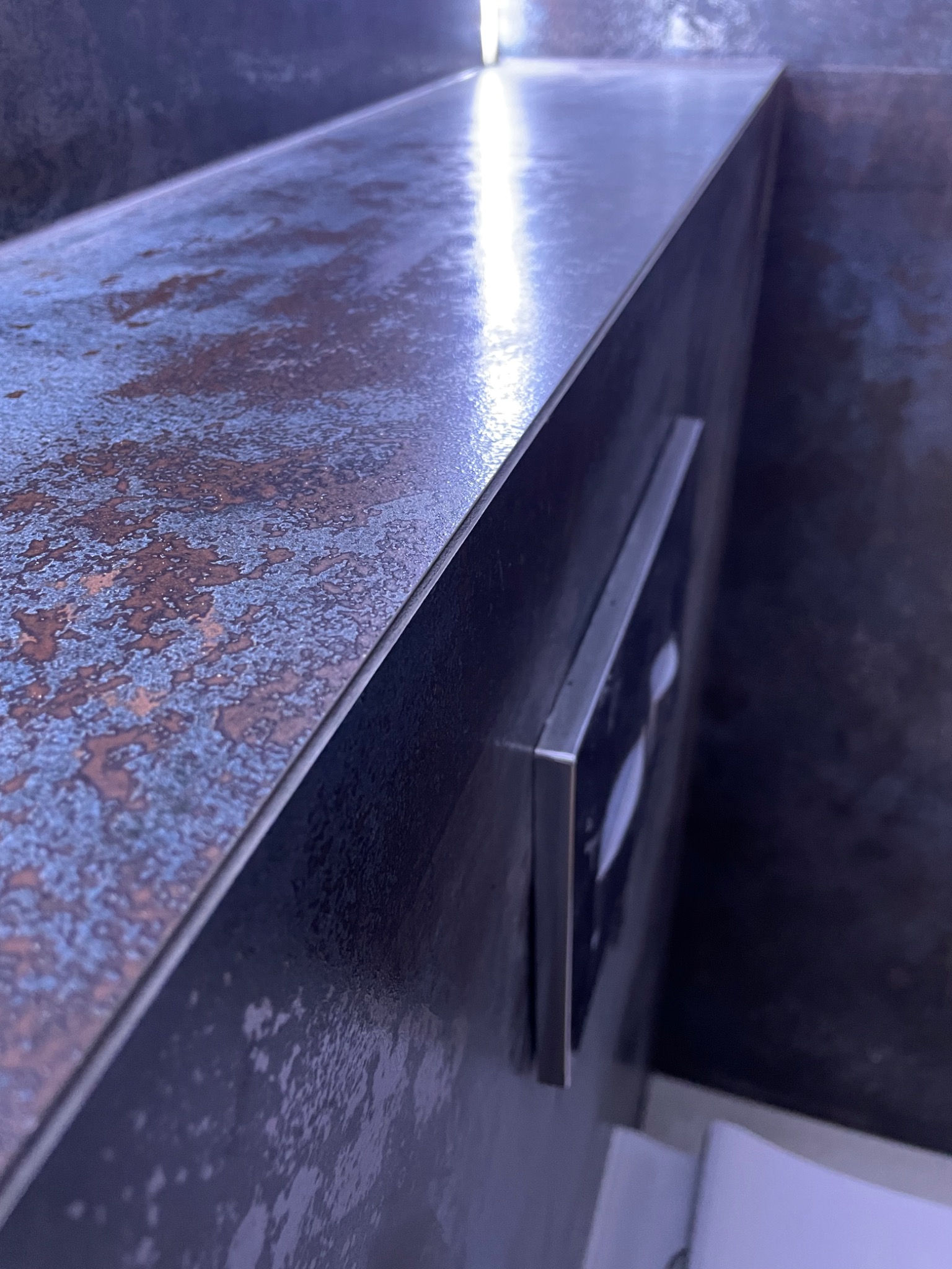 Boczny widok na fragment wykończonej łazienki z umywalką i zabudową, wykończonych płytkami imitującymi rdzę, z widocznym przyciskiem spłuczki.