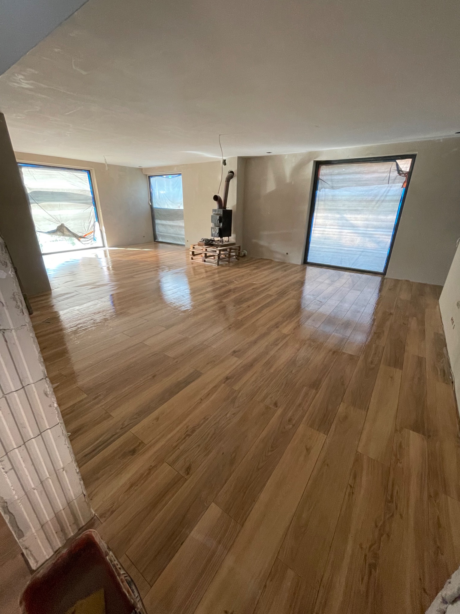 Salon w trakcie prac wykończeniowych: położona drewnopodobna podłoga, okna zabezpieczone folią, ściany po szpachlowaniu, widoczny piec na drewno i wiadro na zaprawę w rogu.