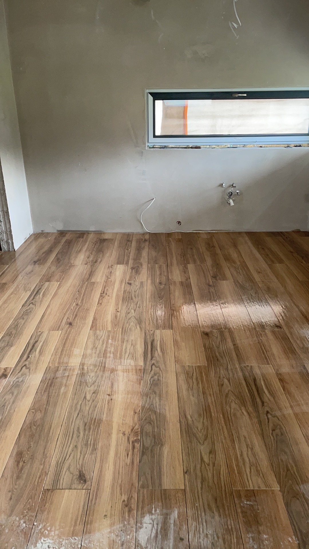 Ułożona podłoga z paneli imitujących drewno w surowym wnętrzu z oknem, ściany przygotowane do malowania, widoczne instalacje hydrauliczne.